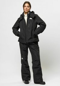 Schwarze wasserdichte Jacke mit Kapuze und Reißverschluss-Taschen; kombiniert mit schwarzen wasserdichten Hosen, die ein weißes Logo tragen. Strukturierter Stoff, lockere Passform.