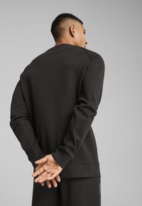 Sweatshirt gri închis cu mâneci lungi și guler rotund. Materialul pare moale și neted. Modelul stă cu mâinile încrucișate în spate.