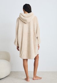 Beige plush hettebadekåpe med myk, teksturert stoff. Har lange ermer og en oversized passform, som slutter midt på låret.