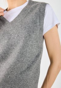 Grå stickad väst med V-ringning, korta ärmar och ribbad nederkant. Materialet verkar mjukt och strukturerat, lagd över en vit t-shirt.