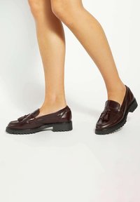 Brown leather loafers with a low heel, featuring a tassel detail, textured sole, and a smooth finish. Suitable for casual wear.