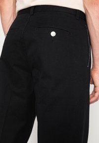 Black cotton trousers with a straight fit, featuring back pockets with silver hardware and white stitching along the side seams.