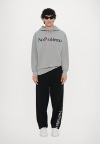 Grauer Hoodie mit dem Text "No Problemo", schwarze Jogginghosen und weiße Sneaker. Das Model trägt schwarze Sonnenbrillen; die Hose hat ein Logo-Detail.