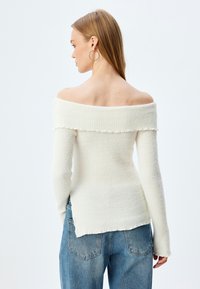 Maglione off-white a spalle scoperte, texture a coste, maniche lunghe, bordi sfrangiati, abbinato a jeans a vita alta blu chiaro.