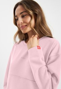 Lichtroze sweatshirt met een ronde halslijn, geribbelde manchetten en een geborduurd logo. Bevat een rode accenttag op de mouw. Zacht materiaal.