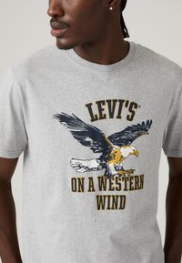 Man in een grijze T-shirt met een afbeelding van een arend in vlucht en de tekst "Levi's On A Western Wind" zichtbaar op de borst.