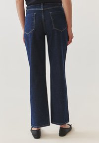 Pantalones vaqueros de mezclilla oscura de pierna ancha con una cintura alta, bolsillos traseros con parche y ligero desvanecimiento. Presentan un dobladillo limpio y costuras resistentes.