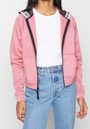 Kvinde iført en lys pink hættetrøje med lynlås og sort kant over en hvid cropped top og højttaljede blå jeans, stående med armene afslappede.