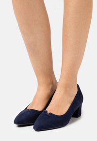 Tacones de ante azul marino con una punta afilada y un pequeño corte en la parte frontal, que presentan un tacón bloque para mayor estabilidad y comodidad.