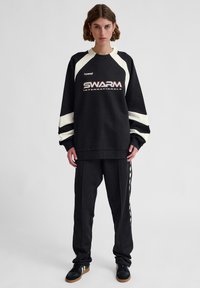 Woman wearing black and white long-sleeve sweatshirt with "SWARM INTERNATIONALS" text, matching black pants, and black sneakers with gum soles.
