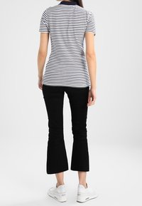 Kobieta ubrana w białą i czarną w prążki krótką koszulę polo, czarne spodnie 3/4 i białe sneakersy, odwrócona plecami na jednolitym tle.