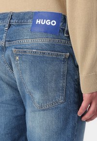 Sininen farkkuhousut, joissa on suorakulmainen sininen etiketti, jossa lukee "HUGO" vyötärönauhan kohdalla, teksturoidulla pinnalla ja korostetulla ompelulla.
