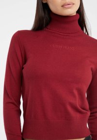 Maglione a collo alto rosso realizzato in tessuto a costine. Presenta il logo ricamato "GUESS JEANS" sul davanti ed è dotato di un design aderente. Maniche lunghe e orlo a costine.