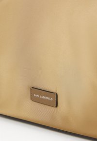 Sac en satin doré avec une texture lisse, orné d'une étiquette rectangulaire marron embossée avec "KARL LAGERFELD". Design minimaliste, sans motifs.