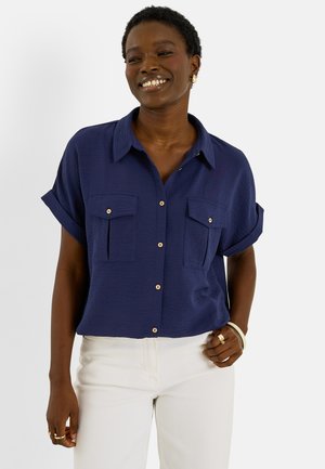 Femme souriante portant une chemise bleu marine à manches courtes boutonnée et un pantalon blanc, avec des boucles d'oreilles et des bagues en or, posant la main sur la hanche.