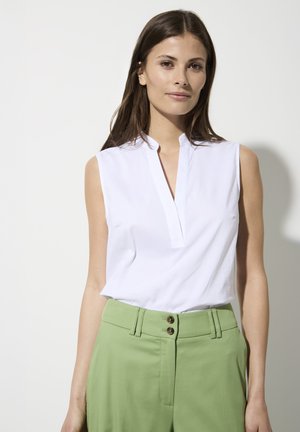 Weiße ärmellose Bluse mit Mandarin-Kragen, kombiniert mit hochgeschnittenen grünen Hosen, die über zwei runde Knöpfe und seitliche Taschen verfügen.