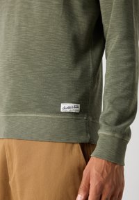 Olijfgroene sweatshirt, gestructureerde stof, ribgebreide manchetten en zoom, met een kledinglabel met "Scotch & Soda" aan de linkerkant.