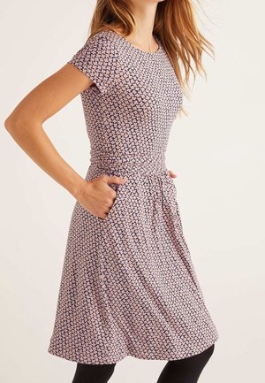 Femme portant une robe à manches courtes et longueur au genou avec un petit motif floral, debout les mains dans les poches de la robe, devant un fond uni.