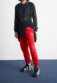 Person som bär en svart Nike-mönstrad långärmad tröja, röda Nike träningsoverallsbyxor, en svart tröja knuten i midjan, och svarta Nike-skor med vit "AIR" text.