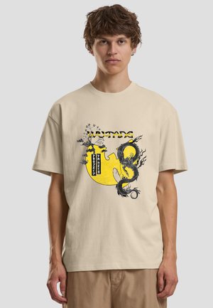 Młody mężczyzna w beżowej koszulce z żółtym logo Wu-Tang, czarnym grafiką smoka i napisem „Project Yureeka” na przodzie.