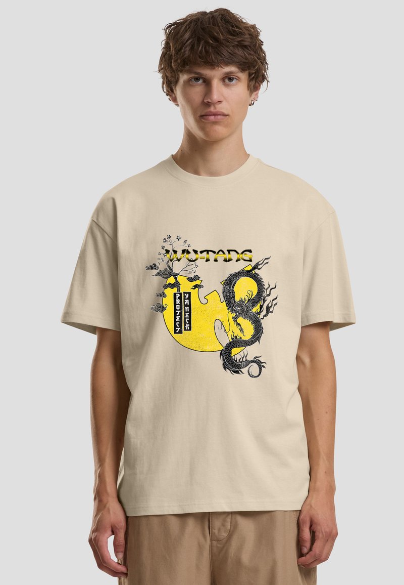 Jeune homme portant un T-shirt beige avec un logo Wu-Tang jaune, un motif de dragon noir, et le texte "Project Yureeka" sur le devant.