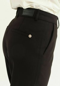 Pantaloni neri sartoriali con una texture liscia, dotati di una tasca posteriore e di un piccolo accento metallico circolare con l'etichetta "FISEAK".