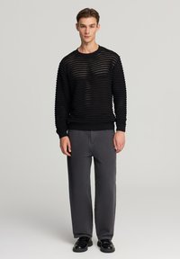 Giovane uomo in piedi con un maglione nero a maglia aperta, pantaloni grigi dritti e scarpe in pelle nere su uno sfondo chiaro e semplice.