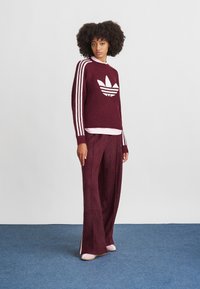 Burgundi kötött pulóver fehér Adidas logóval, három fehér csíkkal a hónaljakon. Széles szárú burgundi textúrázott nadrággal párosítva.