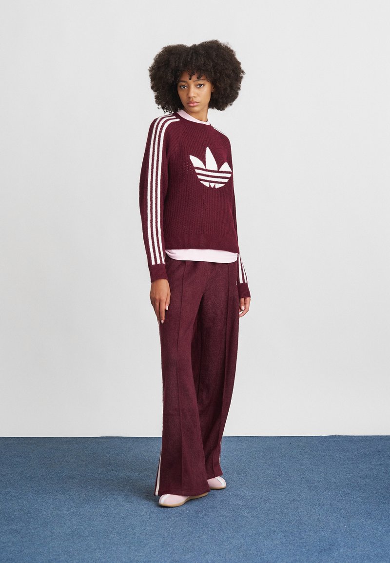 Burgunderfarbener Strickpullover mit weißem Adidas-Logo, der drei weiße Streifen an den Ärmeln hat. Kombiniert mit weiten burgunderfarbenen, strukturierten Hosen.