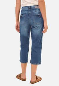 Pantalons capri en denim bleu moyen avec une coupe décontractée, dotés de poches arrière et d'ourlets retroussés, portés avec des sandales brunes à enfiler.
