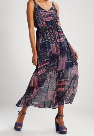 Femme portant une robe maxi sans manches à motif patchwork avec une taille cintrée et des sandales bleues à plateforme à talons hauts.