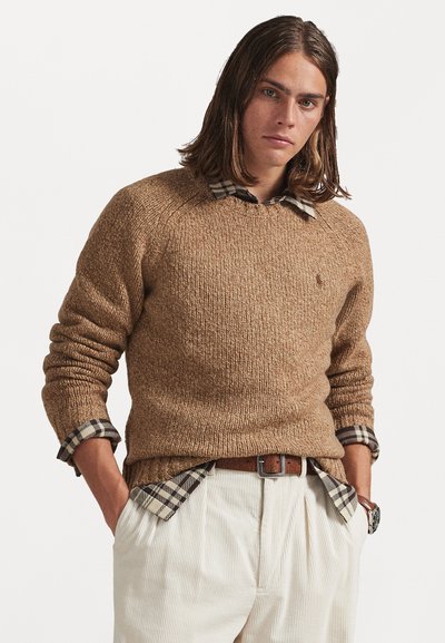 Maglione marrone a maglia con texture a coste, sovrapposto a una camicia a quadri nera e beige; abbinato a pantaloni plissettati color crema.