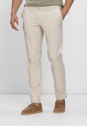 Mann trägt beige Slim-Fit-Hose und braune Wildleder-Schnürschuhe, steht mit einer Hand in der Tasche vor einem schlichten Hintergrund.