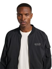 Mand med kort hår iført en sort lynlåstrøje med teksten "HALO" og en hvid skjorte, kigger direkte frem mod en hvid baggrund.