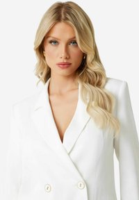 Hvid dobbeltradet blazer med notchet revers, med guldfarvede knapper og en glat tekstur, båret over en v-hals top.