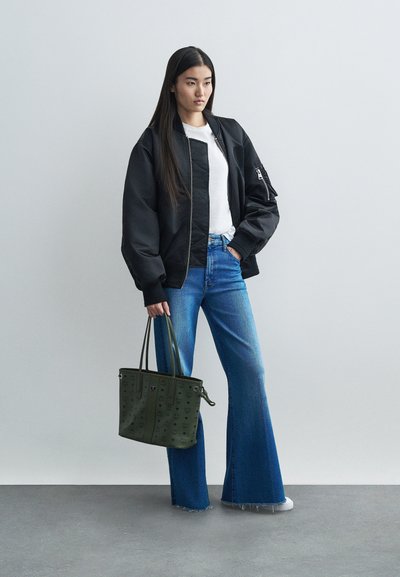 Femeie îmbrăcată cu o jachetă bomber neagră, cămașă albă, blugi evazați albaștri, ținând o geantă de umăr verde măsliniu, stând lângă un perete simplu.