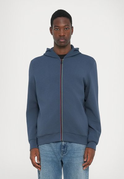 Mørkeblå zip-up hoodie med en kontrasterende flerfarvet lynlås. Blødt materiale, hætte-design og ribbede manchetter. Båret med lyseblå jeans.