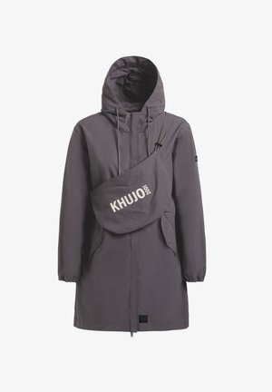 Fiatal nő keresztbe tett lábakkal, szürke kapucnis kabátot, fekete crop topot, fekete leggingseket és fekete masszív csizmát visel.