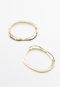 Boucles d'oreilles créoles de couleur or avec un design ondulé, ornées de petites pierres précieuses noires le long d'un bord. Texture métallique lisse, forme moderne.