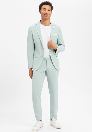 Lächelnder junger Mann, der einen hellgrünen Anzug, ein weißes T-Shirt und weiße Turnschuhe trägt, vor einem einfarbigen weißen Hintergrund stehend.