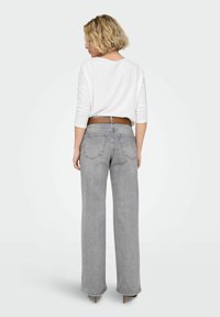 Jeans anchos de pierna en color gris con cintura alta y bolsillos traseros, combinados con una camiseta blanca de manga larga. Cinturón marrón visible en la cintura.