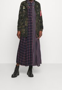 Maskáčová bunda s květinovými detaily přes kostkované košilové šaty v námořnické modři, růžové a bílé, doplněné černými šněrovacími botami.