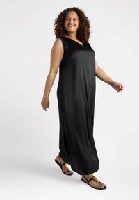 Robe maxi noire sans manches en tissu lisse et brillant avec un décolleté en V et des fentes sur les côtés. Portée avec des sandales noires aux accents dorés.