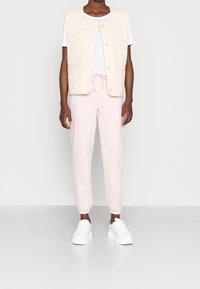 Beige quiltad väst med rund halsringning och knappstängningar, matchad med rosa joggerbyxor och vita sneakers. Slät tygtextur.