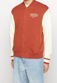 Rost-orange varsityjacka med krämfärgade ärmar, ribbade manschetter, rundad krage, framknappning och Nike-logotypdetalj på bröstet.