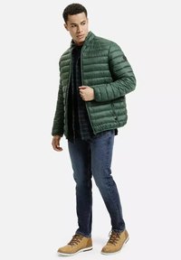 Zielona kurtka puchowa z poziomymi przeszyciami, wysoki kołnierz, boczne zamki, połączona z czarną bluzką, niebieskimi dżinsami i brązowymi butami.