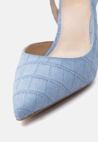 Ανοιχτό μπλε σουέντ παπούτσι με μυτερή μύτη, μοτίβο με ραφές καπιτονέ και λουράκι slingback, εμφανίζεται σε λευκό φόντο.