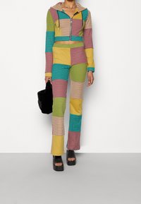 Sweat à capuche court rayé et coloré avec une capuche, accompagné d'un pantalon évasé assorti, présentant un patchwork aux tons verts, jaunes, roses et bleus. Sac noir inclus.
