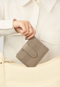 Grijze leren portemonnee met kaartenhouders, vastgehouden door een hand, gedeeltelijk in een crèmekleurige leren tote bag. Heeft een gladde textuur en een drukknoopsluiting.