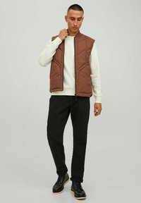 Brown quilted vest with zip closure, featuring vertical seams and two side pockets, worn over a white sweater and paired with black pants and boots.